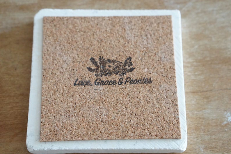 May include: Square coaster with a white border and a cork backing. The cork backing has a floral design and the text "Love, Grace & Peonies" printed in black. The coaster is likely made of ceramic or stone.