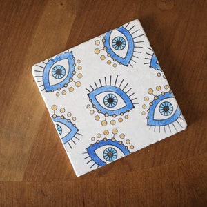 May include: A square white ceramic coaster featuring a repeating pattern of blue and black evil eye motifs. Each eye design includes long black eyelashes and small yellow circular accents surrounding the iris, creating a consistent graphic print.