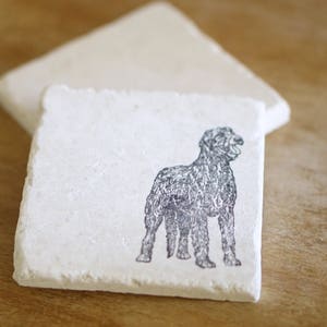 May include: Square, off-white stone coasters. One coaster features a black illustration of a dog. The dog is standing with its mouth open. A second coaster is behind the first. The coasters are on a wooden surface.