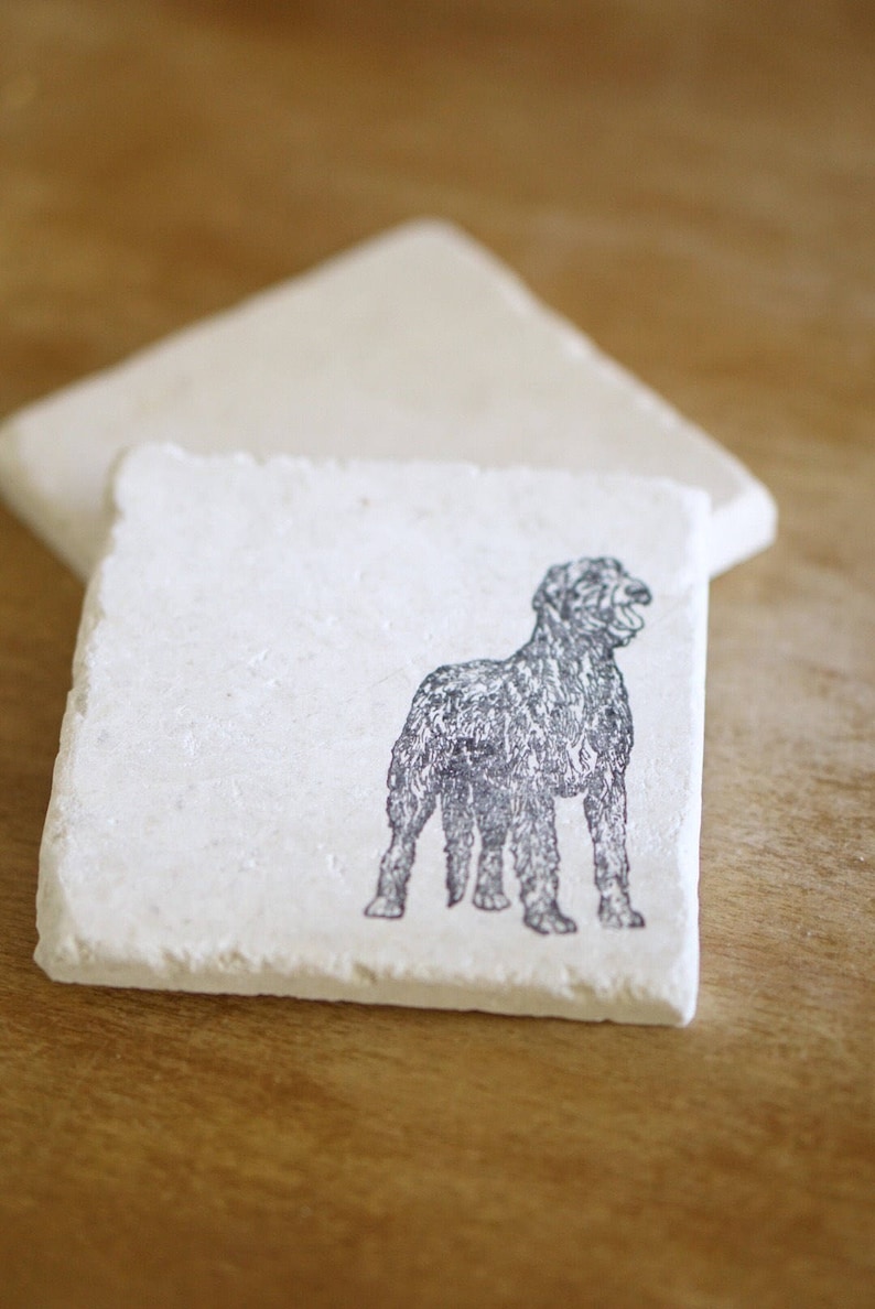 May include: Square, off-white stone coasters. One coaster features a black illustration of a dog. The dog is standing with its mouth open. A second coaster is behind the first. The coasters are on a wooden surface.