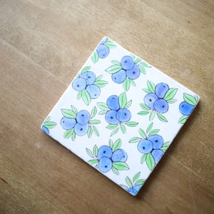 May include: A square ceramic coaster featuring a repeating pattern of hand-painted blue blueberries with green leaves on a white background. The design displays multiple clusters of berries arranged across the surface of the tile.