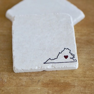May include: Two white square coasters with a black outline of the state of Virginia and a red heart in the center. The coasters are on a wooden surface.
