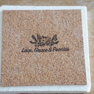 May include: A white ceramic coaster with a cork top. The cork top has a floral design and the text "Love, Grace & Peonies".