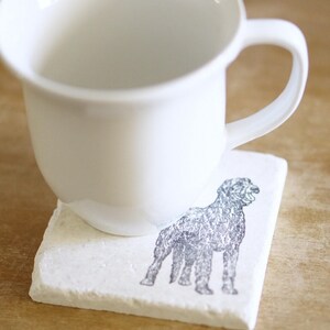 May include: A white ceramic mug rests on a square, off-white coaster. The coaster features a detailed illustration of a dog in shades of gray. The mug has a simple, rounded design with a curved handle.