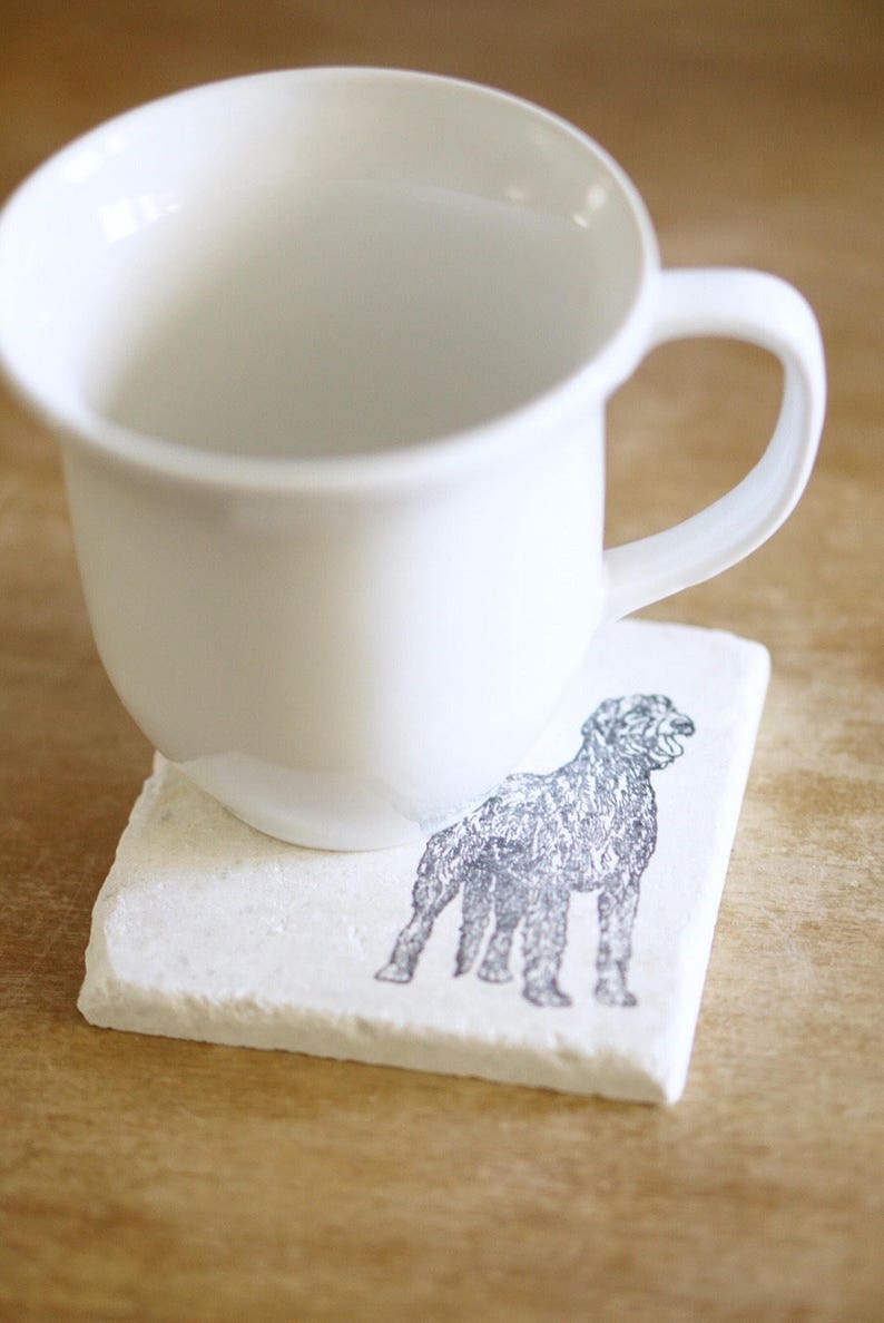 May include: A white ceramic mug rests on a square, off-white coaster. The coaster features a detailed illustration of a dog in shades of gray. The mug has a simple, rounded design with a curved handle.
