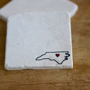 May include: White ceramic coaster with a black outline of the state of North Carolina and a red heart in the center.