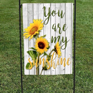 May include: A white wooden sign with a rustic look featuring three yellow sunflowers and the text "You are my sunshine". The sign is displayed on a black metal garden stake.