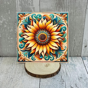 May include: Square decorative tile featuring a vibrant sunflower design. The sunflower has yellow and orange petals with a detailed brown center. Teal and orange decorative elements frame the flower. The tile is displayed on a wooden slice.