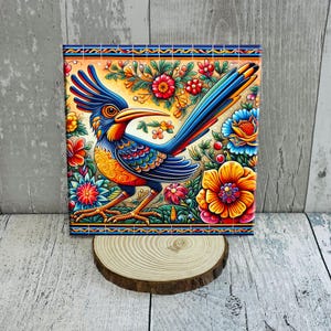 May include: A square decorative tile featuring a colorful illustration of a roadrunner bird. The bird has blue, orange, and yellow feathers, surrounded by vibrant flowers. The tile is set on a wooden slice, with a decorative border.