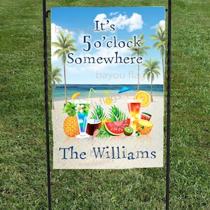 May include: A garden flag with a beach scene, palm trees, and cocktails. The text reads "It's 5 o'clock Somewhere" and "The Williams". The flag is mounted on a black metal stand and set in green grass.