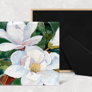May include: Square art print featuring a watercolor painting of white magnolia flowers with yellow centers and green leaves. The artwork is displayed on a wooden easel with a black backing.