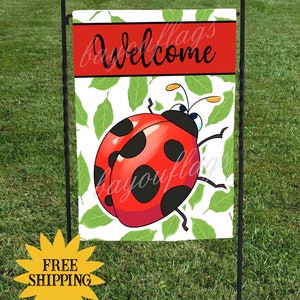May include: A welcome garden flag with a red ladybug on a white background with green leaves. The flag has the word "Welcome" in black text on a red banner at the top. The flag is attached to a black metal garden flag stand.