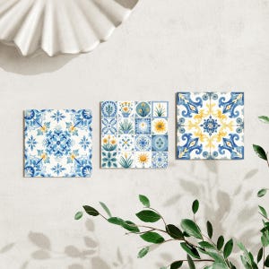 Könnte beinhalten: Drei quadratische Keramikfliesen mit blau-gelben Blumenmustern. Die Fliesen weisen verschiedene Designs auf, darunter stilisierte Blumen, geometrische Formen und Sonnenmotive. Die Fliesen sind vor einem weißen Hintergrund mit einer dekorativen Muschel und Grün angeordnet.