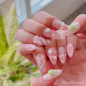 Op de afbeelding: Roze en witte nagelkunst met verschillende ontwerpen, waaronder strikken, harten, bloemen en glitter. De nagels zijn lang en amandelvormig. De tekst "tomatoesnails" is zichtbaar in de rechterbenedenhoek van de afbeelding.