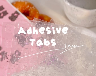 Può includere: Primo piano di un pacchetto di adesivi trasparenti. Il pacchetto è etichettato "Adhesive Tabs" e "1 piece".