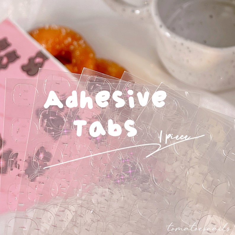 May include: A close-up of a package of clear adhesive tabs. The package is labeled "Adhesive Tabs" and "1 piece".