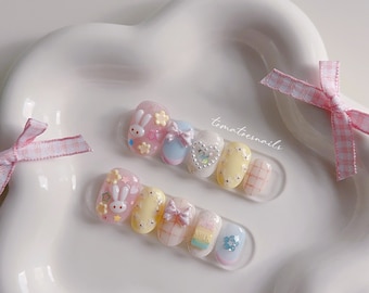 May include: A set of pastel-colored press-on nails with cute bunny and bow designs. The nails are decorated with glitter, rhinestones, and small flowers. The set includes a variety of shapes and sizes.