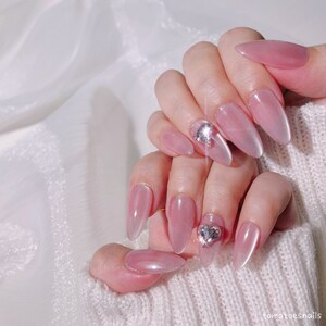 May include: Pink almond-shaped nails with a clear, iridescent finish. Some nails have a small, clear rhinestone heart accent.