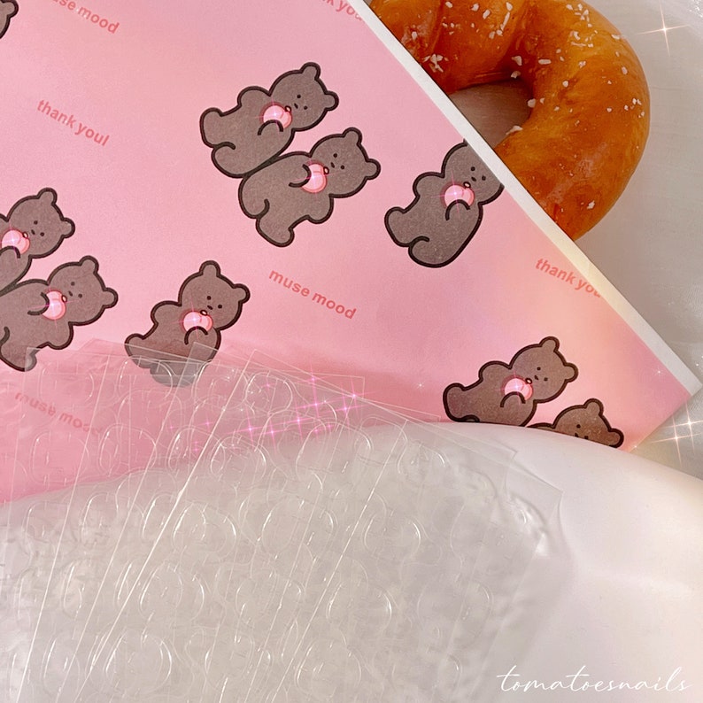 May include: A pink sheet of paper with a repeating pattern of brown bears holding pink hearts. The text "muse mood" and "thank you" is printed on the paper. A clear plastic sheet with multiple small circles is on top of the paper.
