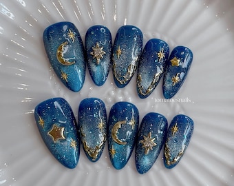 May include: A set of ten blue and glittery artificial nails. Each nail features celestial designs, including gold moons, stars, and decorative accents. The nails are arranged on a white plate. The text "tomatoesnails" is visible.
