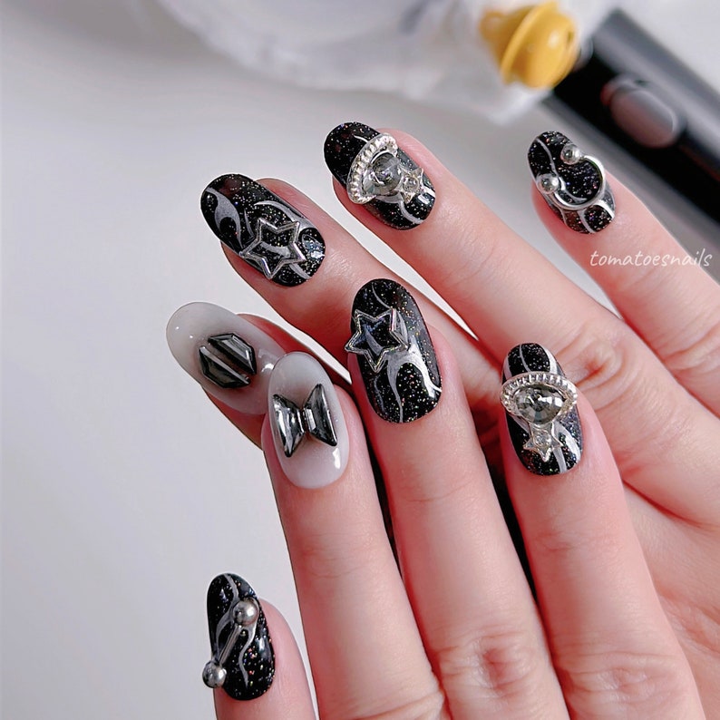 May include: Close-up of a hand with black and silver nail art featuring stars, bows, and rings. The nails are painted black with silver glitter and have various silver embellishments.