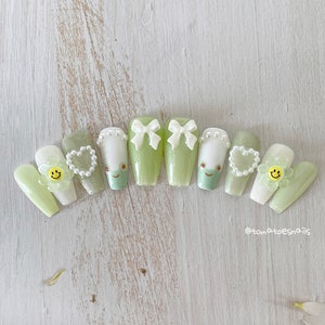 May include: A set of ten green and white nail tips with various decorations, including bows, hearts, and smiley faces. The nail tips are arranged in a semi-circle on a white wooden surface. The text "@tomatoesnails" is visible in the bottom right corner of the image.