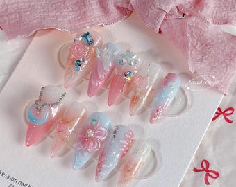Può includere: Una collezione di unghie finte con vari design. Ogni unghia presenta un design unico, tra cui vortici rosa e blu, accenti floreali e abbellimenti scintillanti. Alcune unghie hanno un dettaglio a catena. Le unghie sono disposte su una superficie bianca.