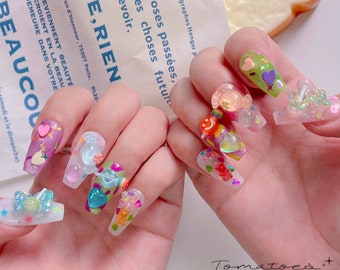 May include: Close-up of a hand with colourful, decorated nails. The nails are long and have a variety of 3D embellishments, including hearts, candy, and glitter. The text "Tomatoest nails + new in stock" is visible at the bottom of the image.