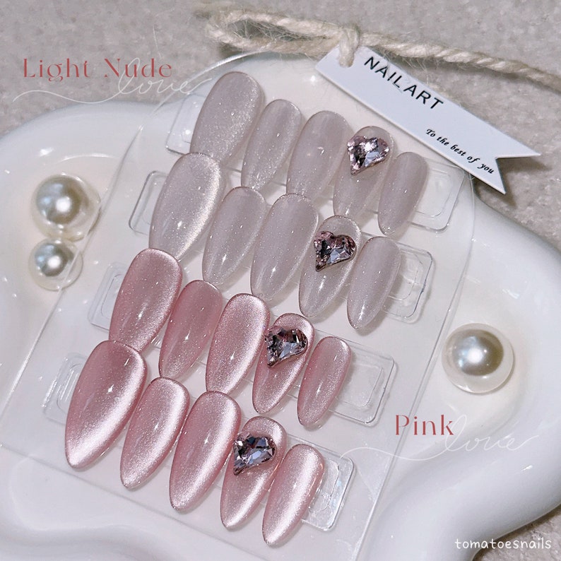 May include: Two sets of false nails with a heart-shaped rhinestone on each nail. The top set is light nude and the bottom set is pink. The text "NAIL ART" and "To the best of you" is on a white tag. The text "Light Nude" and "Pink" is on the bottom of the image.