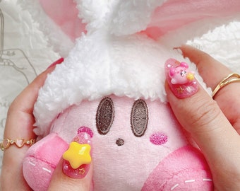 May include: Pink and white plush toy with a pink face and brown eyes. The toy is being held by a hand with pink and yellow nail art featuring a star and a cartoon character. The nail art is sparkly and has glitter.