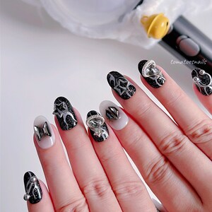 May include: Close-up of a hand with black and silver nail art. The nails are oval shaped and have a black base with silver glitter and various silver embellishments. The nail art features stars, bows, and other decorative elements.