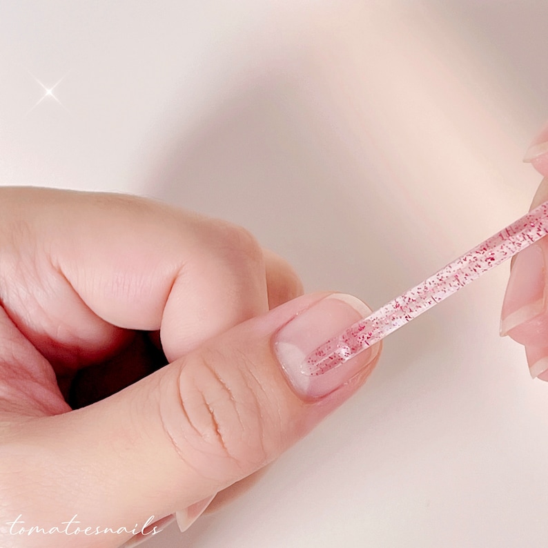 Puede incluir: Una herramienta de u&ntilde;as transparente con un acabado de purpurina rosa se utiliza para suavizar una aplicaci&oacute;n de esmalte de u&ntilde;as rosa.