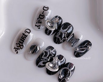 Puede incluir: Conjunto de seis uñas postizas negras y blancas con detalles plateados. Las uñas presentan una variedad de diseños, incluyendo estrellas, remolinos y pedrería. Las uñas también tienen texto, incluyendo "D-Day" y "August".
