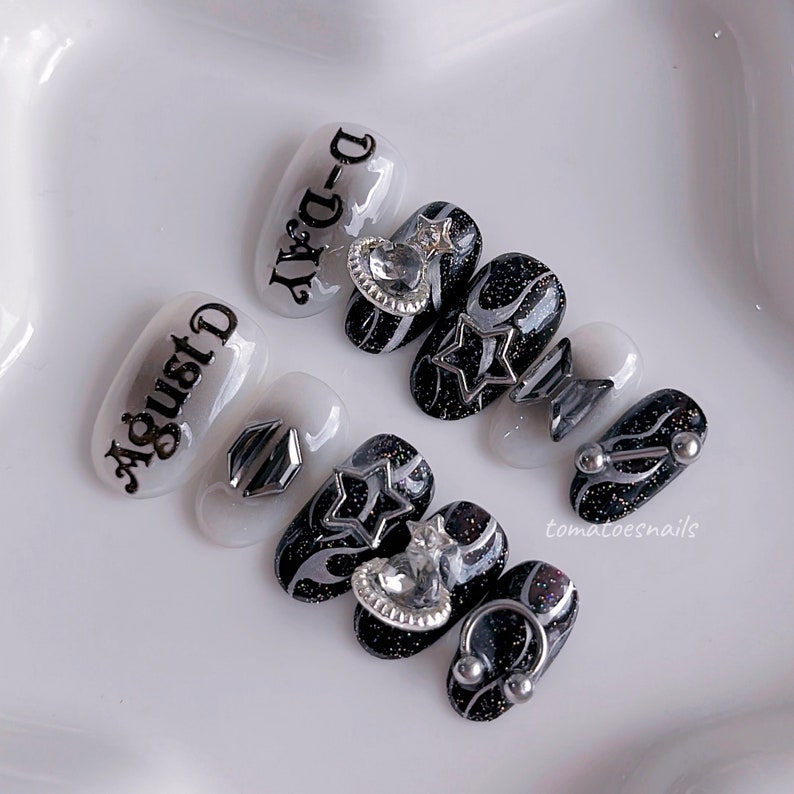May include: Set of six black and white press-on nails with silver accents. The nails feature a variety of designs, including stars, swirls, and rhinestones. The nails also have text on them, including "D-Day" and "August".