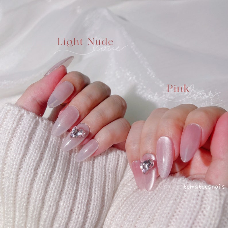 May include: Two hands with long, almond-shaped nails. The nails are painted in two different colors: light nude and pink. Each nail has a small, clear rhinestone on it. The text "Light Nude" and "Pink" are written above the nails.