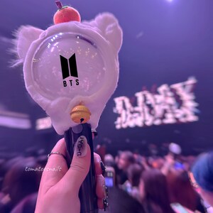 May include: A white plush light stick with a clear plastic sphere containing the letters "BTS" in black. The light stick has a yellow bell and a black handle with the text "D-DAY" in black.