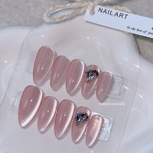 May include: Set of ten pink oval-shaped press-on nails with a sparkly finish. Two nails feature a clear heart-shaped rhinestone.