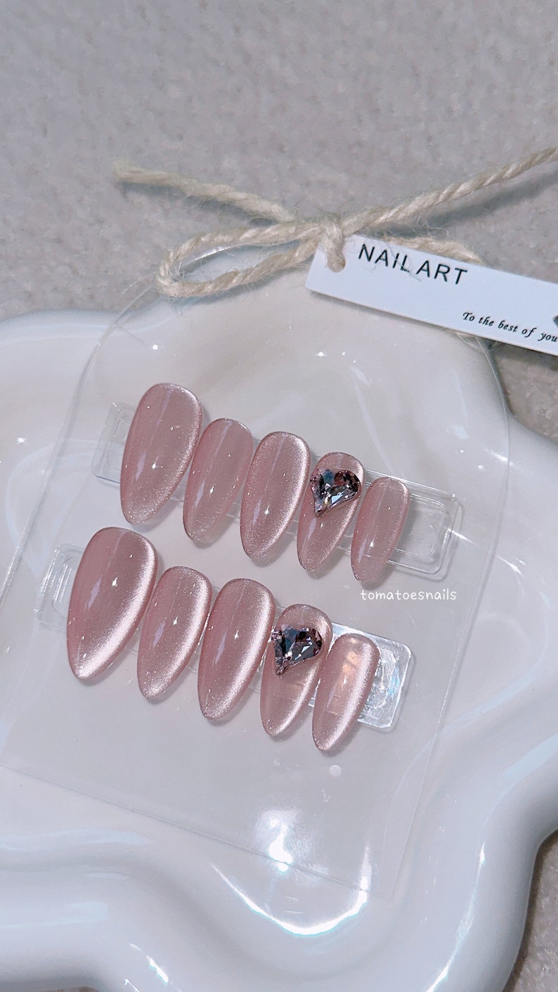 May include: Set of ten pink oval-shaped press-on nails with a sparkly finish. Two nails feature a clear heart-shaped rhinestone.