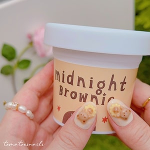 May include: A small container with a white lid and a label reading "midnight Brownie". The container is held by a person with decorated nails featuring bear and sunflower designs. A pearl ring is also visible.