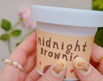 May include: A small, light-colored container labeled "midnight Brownie" with a white lid. The container is held by a person with decorated nails featuring bear and sunflower designs. A pearl ring is also visible.