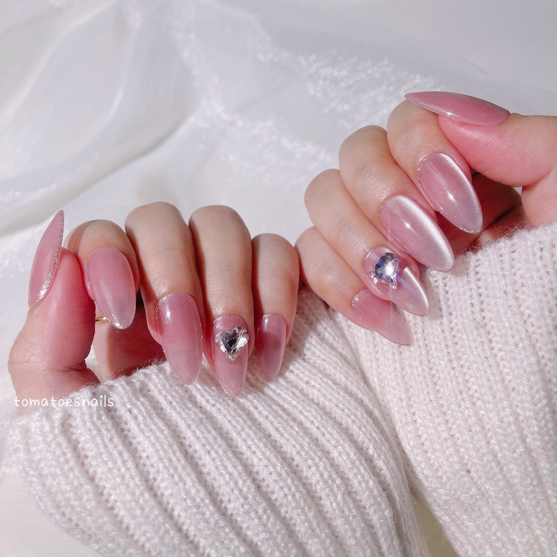 May include: Close-up of a hand with pink almond-shaped nails with a clear, iridescent finish. Some nails have small, clear rhinestones.