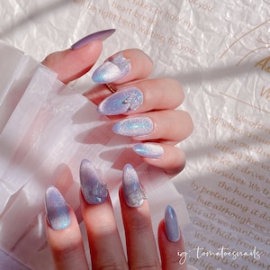 May include: Close-up of a hand with long, oval-shaped nails painted a light purple with a shimmery finish. Each nail is adorned with a small, 3D butterfly charm.