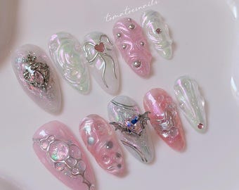 May include: A set of 10 pink and white iridescent press-on nails with various silver and black embellishments, including a heart, a bat, and a crown. The nails are decorated with swirls and glitter.