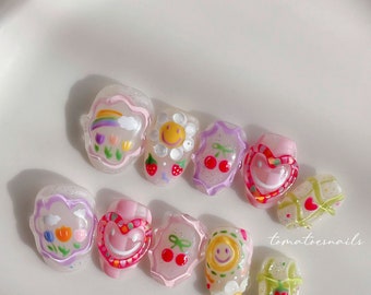 May include: A set of 10 colorful nail art designs featuring a rainbow, smiley faces, cherries, hearts, and flowers. The designs are on a clear background with glitter accents. The text "tomatoenails" is visible in the bottom right corner of the image.