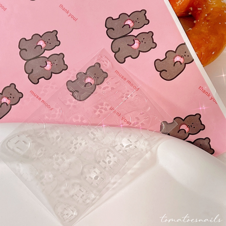 May include: A clear plastic sheet with multiple small circles, designed for nail art. The sheet is partially covering a pink background with a repeating pattern of brown bears holding pink donuts. The text "thank you!" and "muse mood" are visible on the background.