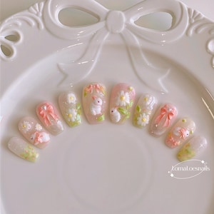 May include: A set of ten white oval-shaped false nails with 3D bunny and flower designs. The nails are decorated with pink bows, white bunnies, and white flowers with green leaves. The nails are arranged in a semi-circle on a white plate. The text "tomatoesnails" is visible in the bottom right corner of the image.