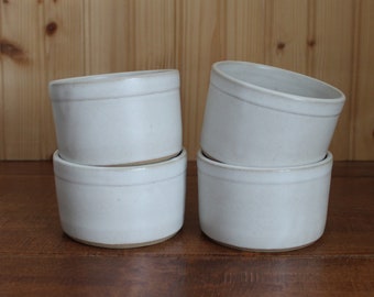 May include: Four white ceramic ramekins are stacked on a wooden surface. The ramekins are cylindrical with a slightly flared rim and a subtle, light-colored band around the middle. The background is a wooden wall.