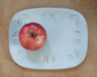 May include: A white ceramic serving platter with a raised leaf design around the edge. A red apple sits in the center of the platter.