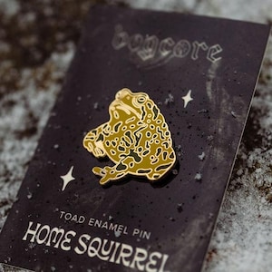 May include: A gold and green toad enamel pin on a black card with the text "bogcore" and "HOME SQUIRREL". The pin is shaped like a toad with a detailed pattern. The card also has the text "TOAD ENAMEL PIN".
