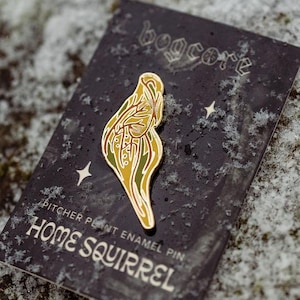 Könnte beinhalten: Ein goldfarbener Emaille-Pin in Form einer Kannenpflanze mit grünen und gelben Details wird auf einer schwarzen Karte präsentiert. Auf der Karte steht "HOME SQUIRREL" und "PITCHER PLANT ENAMEL PIN". Der Hintergrund ist eine schneebedeckte Oberfläche.